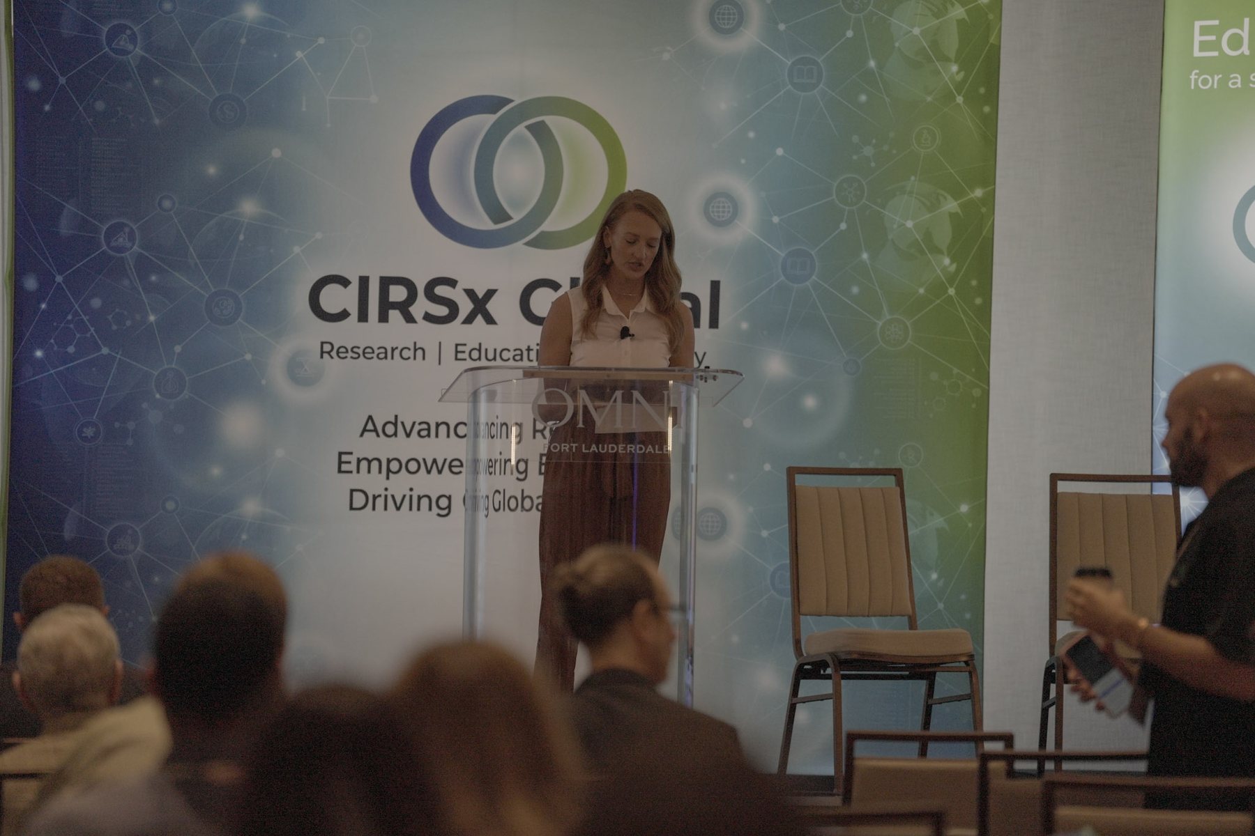 A wide view of a conference room, a speaker presenting beside the event banner