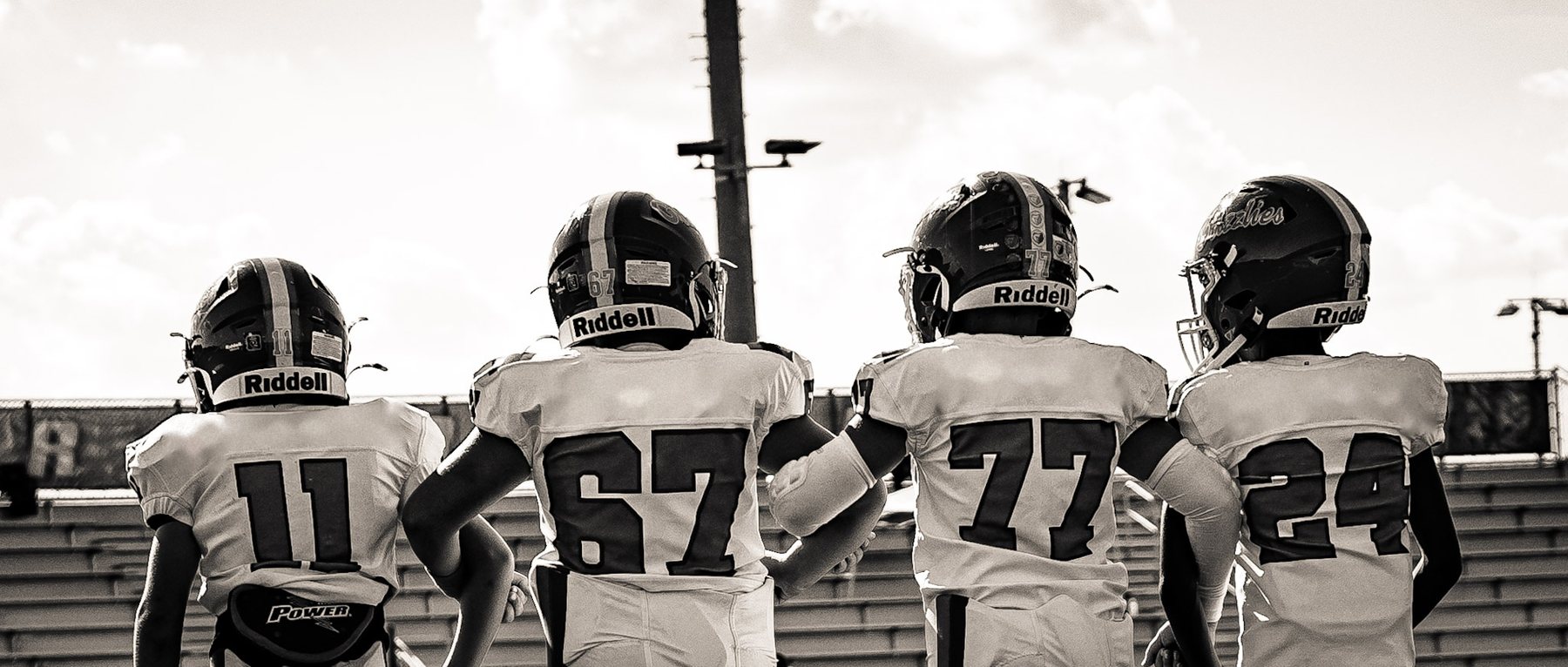 Black-and-white photo of youth football players seen from behind, helmets on, arms around each other