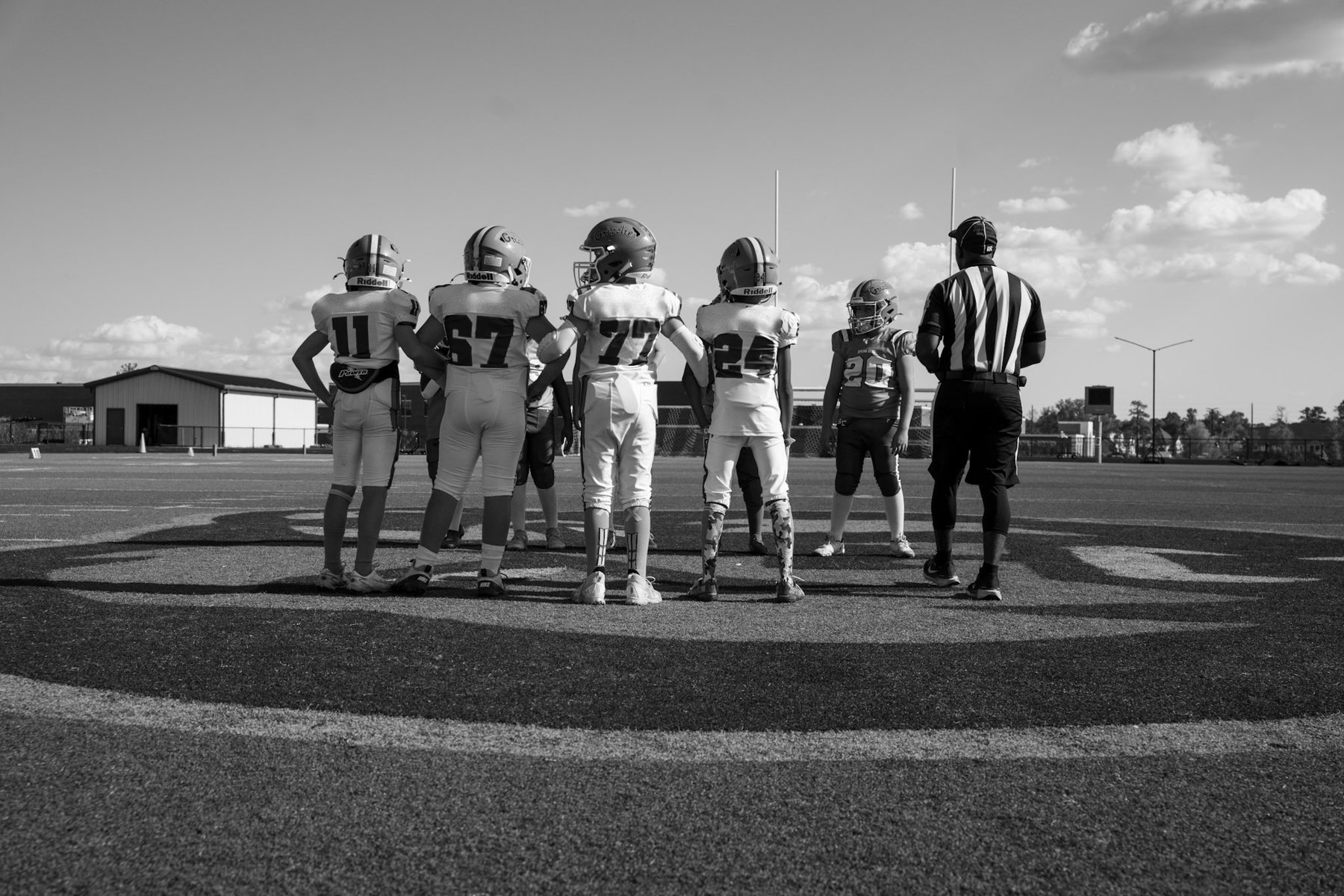 Black-and-white photo of youth football captains and a referee at midfield for the coin toss, seen from behind