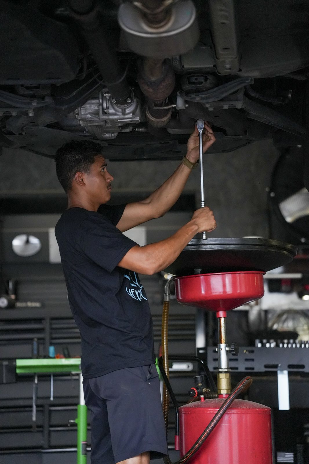 A mechanic working overhead with a long ratchet above a red oil drain